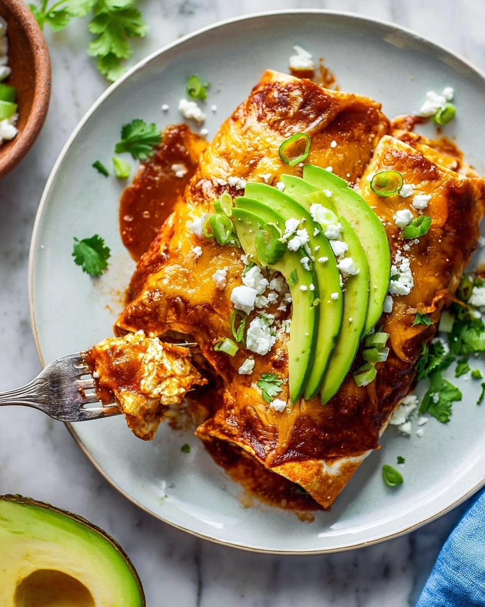 A close-up view of two layered enchiladas on a white plate shows the bottom layer of soft tortillas soaked in red sauce, topped with melted golden and light brown cheese and a rich reddish-brown sauce covering the edges. On top, two bright green slices of fresh avocado are placed, sprinkled with small white crumbles of cheese and tiny rings of light green scallions. Sprigs of fresh cilantro add a touch of dark green on and around the enchiladas. A silver fork lifts a piece from the left side, showing the soft, saucy texture inside. The plate sits on a white marbled surface with part of a halved avocado and a small clay bowl visible nearby. photo taken with an iphone --ar 4:5 --v 7