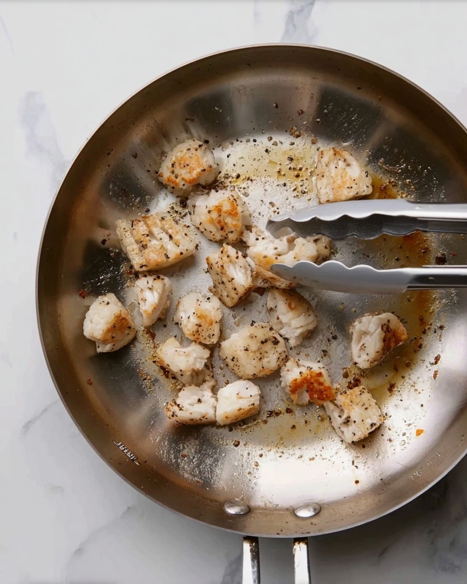 A close-up top view of a silver pan sitting on a white marbled surface, with small pieces of cooked food inside that are light brown and white, some pieces with black pepper spots. A pair of silver tongs is holding a few pieces above the pan. The pan has some browned spots and oil stains, showing that the food is being cooked. Photo taken with an iphone --ar 4:5 --v 7