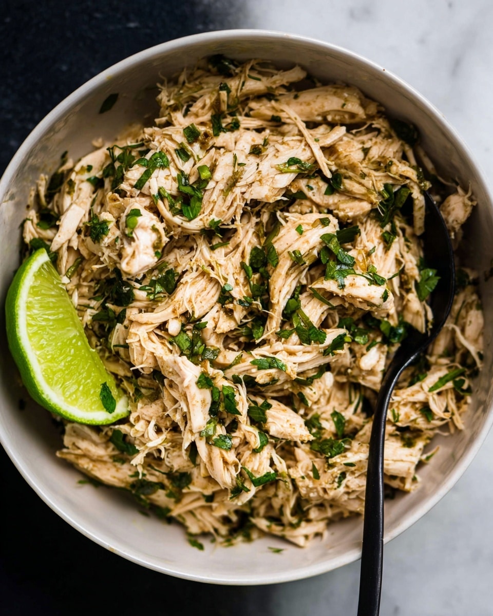 A close-up view of shredded cooked chicken mixed with small pieces of chopped green herbs, all served in a deep white bowl. The chicken has a light brown color with a slightly moist texture, and the herbs are scattered evenly, adding a fresh green contrast. A wedge of bright green lime rests on one side of the bowl. A black spoon is placed inside the bowl, partially submerged in the chicken mixture. The bowl sits on a white marbled surface. photo taken with an iphone --ar 4:5 --v 7