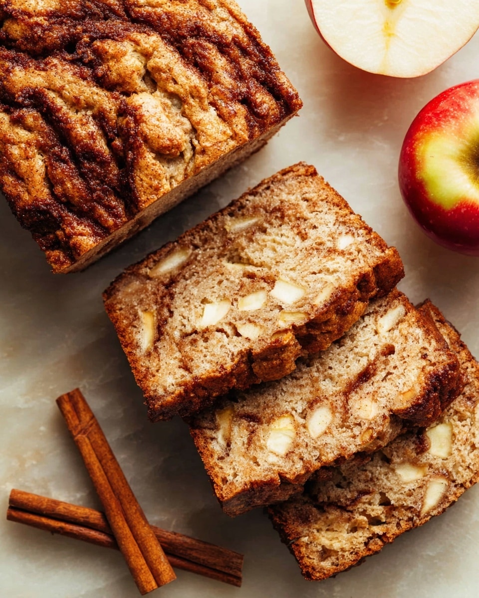 The image shows a loaf of apple cinnamon bread with three slices cut and laid next to it. The bread has a golden-brown crust with a textured top layer showing swirls of dark cinnamon. Inside, the bread is light brown, soft, and full of small apple pieces scattered throughout. The bread rests on a white marbled surface near three sticks of cinnamon and a red and yellow apple on the right side. The overall look is warm and inviting, with a mix of soft and crumbly textures. photo taken with an iphone --ar 4:5 --v 7
