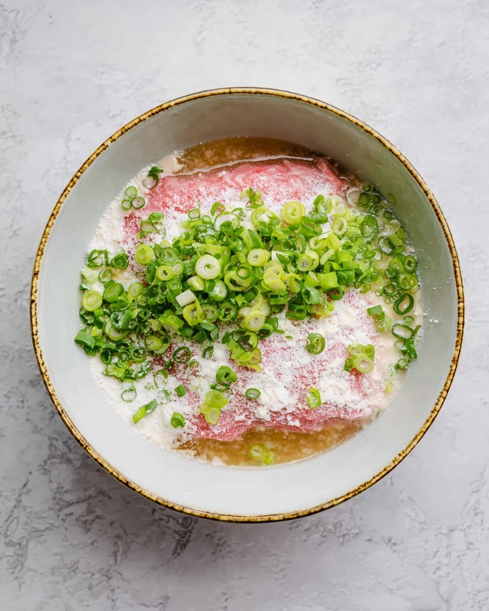 A white bowl with a gold rim sits on a white marbled surface, holding a mix of three layers: the bottom layer is a light brown liquid sauce, the middle layer is a smooth pink raw meat block, and the top layer consists of white powder sprinkled over the meat with many bright green sliced scallions spread unevenly on top. The overall look is fresh and colorful with the contrast between the pink, green, and white photo taken with an iphone --ar 4:5 --v 7