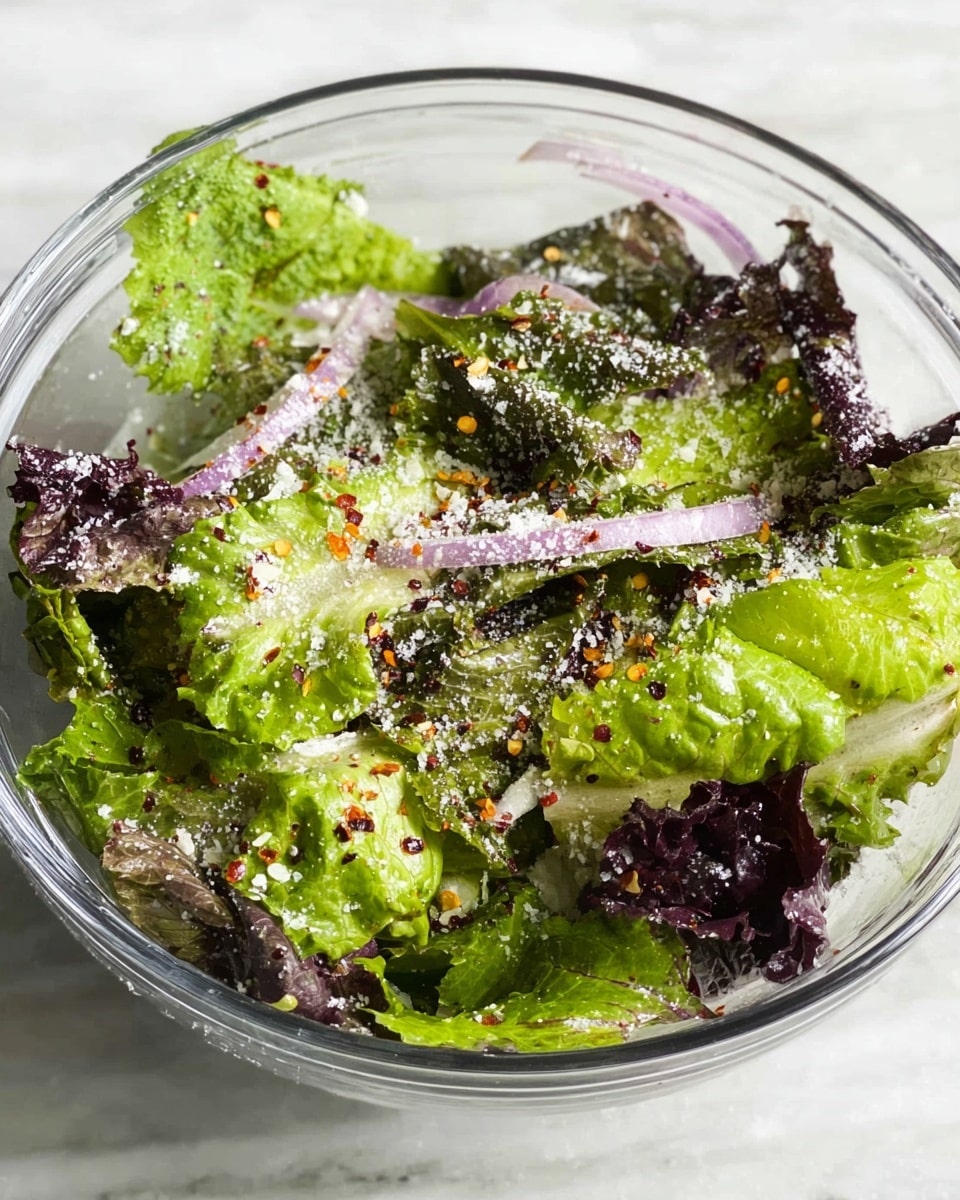 A clear glass bowl filled with mixed green salad leaves showing different shades of green and purple, layered with thin slices of light purple onion rings spread on top. The salad is sprinkled with white grated cheese and small red chili flakes that add specks of red and yellow across the leafy greens. The bowl is placed on a white marbled texture surface, adding a clean and fresh look. Photo taken with an iphone --ar 4:5 --v 7