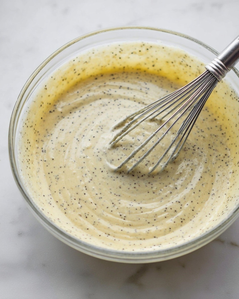 A clear glass bowl filled with a pale yellow batter speckled with tiny black poppy seeds, showing a smooth and creamy texture with some gentle swirls from mixing. A metal whisk rests inside the bowl, partly covered by the batter, with the whisk wires thinly coated in the same speckled mixture. The bowl is placed on a white marbled surface, and the image captures a close-up view focusing on the batter and whisk. photo taken with an iphone --ar 4:5 --v 7