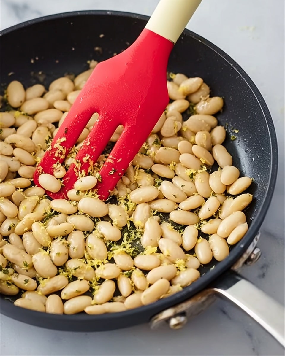 A close-up view of a black frying pan filled with light beige beans mixed with small bits of yellow zest and sprinkled with green herb flakes, softly stirred by a red silicone spatula with a cream handle. The pan sits on a white marbled surface with two metal rivets visible on its edge. The beans are plump and smooth, filling most of the pan, while the spatula rests on the top right side of the pan, partly in contact with the beans. Photo taken with an iphone --ar 4:5 --v 7