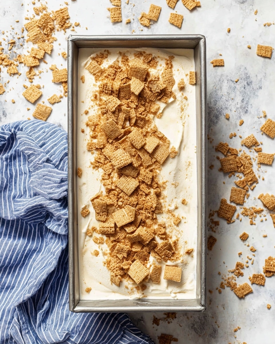 A white bowl holds three scoops of creamy beige ice cream with small specks, topped with crumbly golden brown pieces, some melting slightly, giving a soft, smooth texture. The bowl sits inside another white bowl, both placed on a white marbled surface with light brown crumbs scattered around. A slim golden spoon is placed inside the bowl, leaning against its side. In the softly blurred background, a blue and white striped cloth adds a cozy touch. photo taken with an iphone --ar 4:5 --v 7