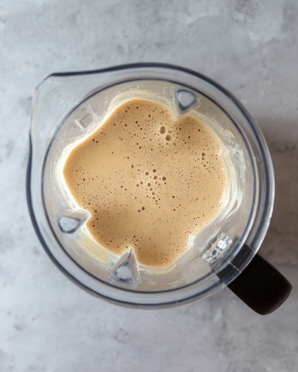 The image shows a blender jar filled with a light tan, creamy liquid that has small bubbles on the surface. The blender jar is clear with a black handle on the right side and is placed on a white marbled texture. The creamy liquid inside fills about half of the jar, and the top surface is smooth with tiny bubbles spread evenly. photo taken with an iphone --ar 4:5 --v 7