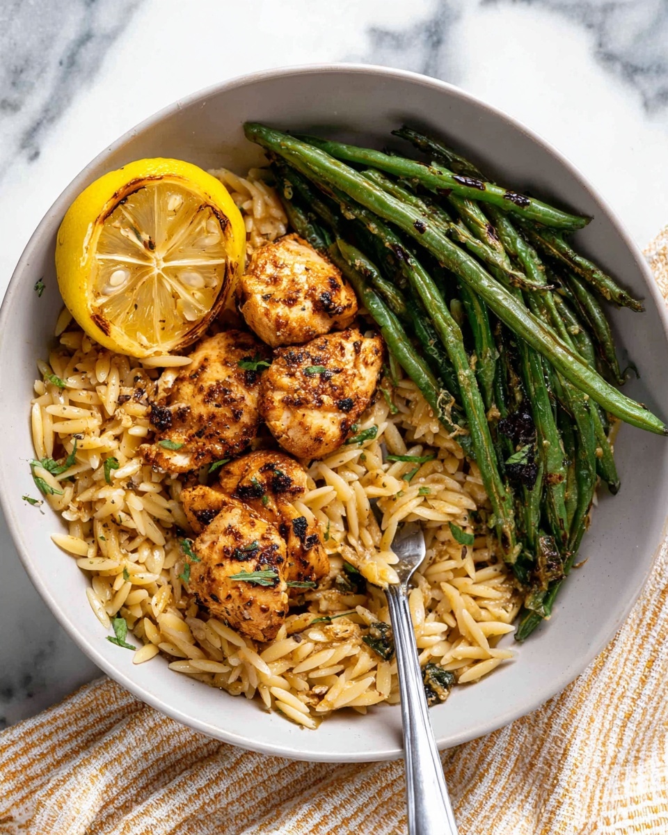 A white bowl holds three main layers: the bottom layer is a light brown orzo pasta mixed with small green herbs, the middle layer features round, golden-brown seasoned chicken pieces with visible black spices, and a charred yellow lemon slice on the left side, while the top right side has a neat pile of shiny, grilled green beans with dark grill marks. A silver fork rests diagonally across the orzo and chicken, partly buried. The bowl sits on a white marbled surface with a beige and white striped cloth underneath. Photo taken with an iphone --ar 4:5 --v 7