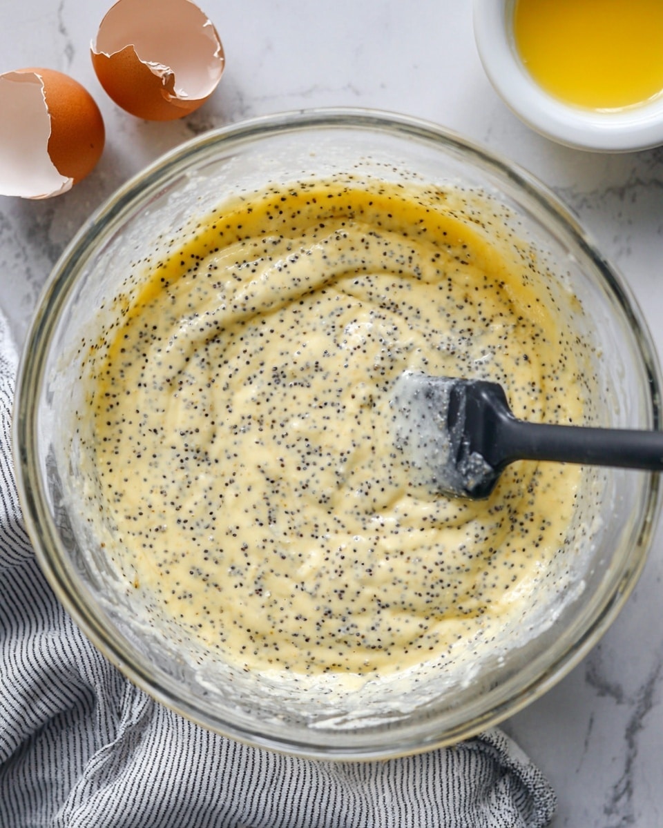 A clear glass bowl holds a thick, creamy yellow batter mixed with many small black poppy seeds spread evenly throughout. A black spatula with some batter on its edge rests inside the bowl, partially submerged in the mixture. Behind the bowl are two cracked brown eggshells on a white marbled surface, and a white bowl with a small amount of yellow liquid is visible in the lower right corner. A striped cloth is partially visible under the bowl, adding texture to the scene. Photo taken with an iphone --ar 4:5 --v 7