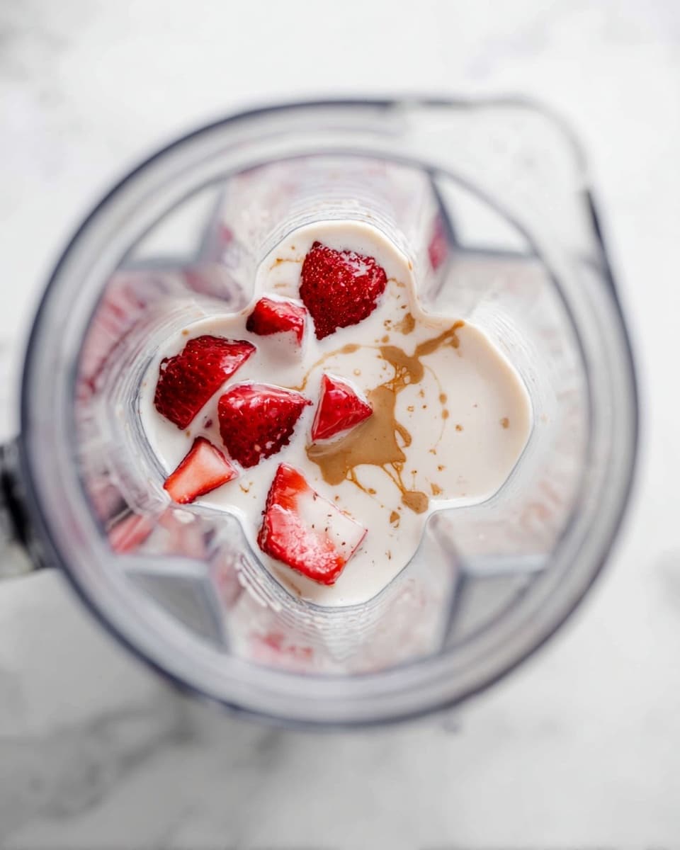 The image shows a blender from above with its clear container filled with several ingredients. At the bottom, there are pieces of red strawberries, some partially covered in a white creamy liquid. The creamy liquid layers around and on top of the strawberries, with a drizzle of light brown vanilla or syrup on the surface, slightly mixing into the cream. The blender is placed on a white marbled surface. The photo taken with an iphone --ar 4:5 --v 7