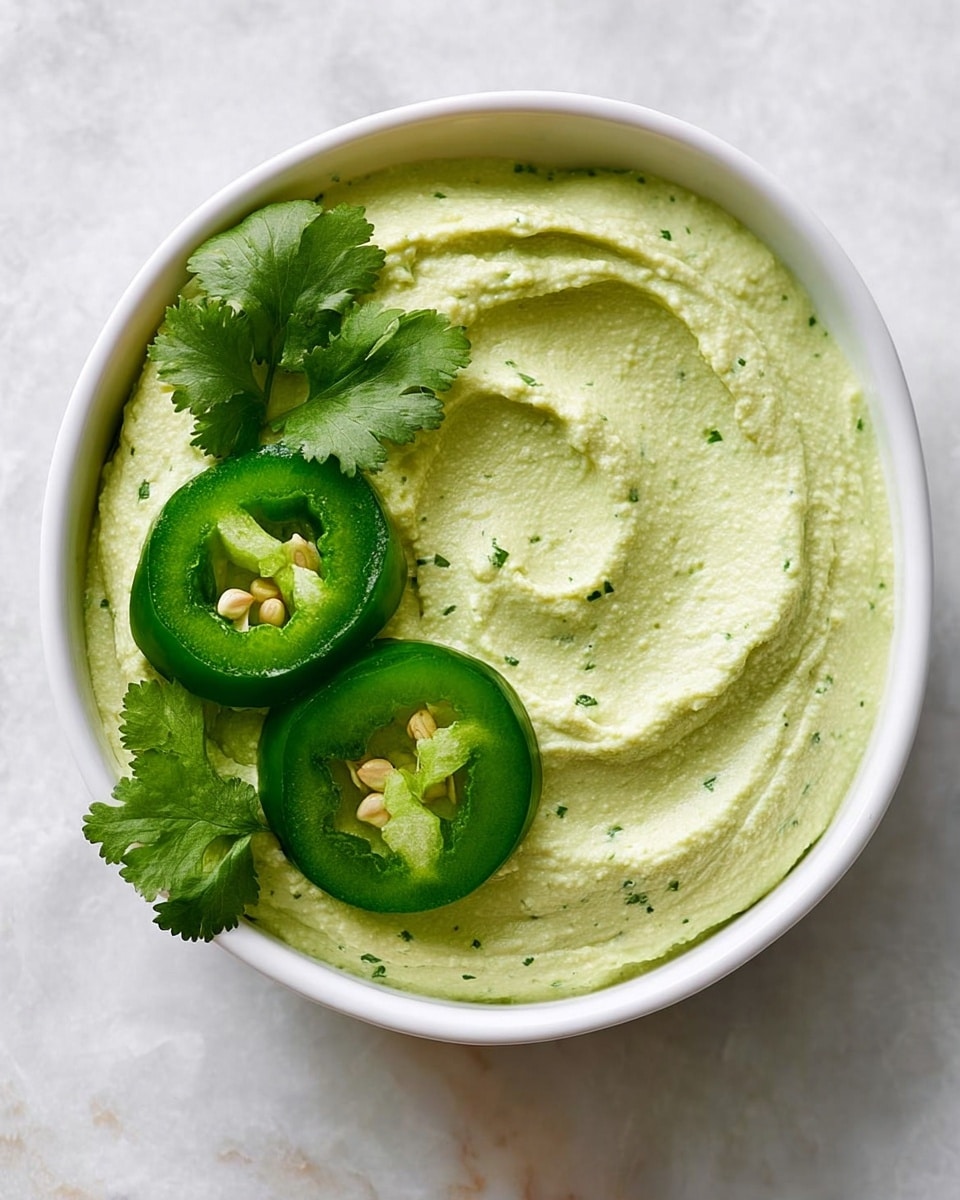 A white bowl holds a smooth, creamy pale green dip with subtle specks of herbs, spread evenly with soft swirls visible on the surface. On top, three slices of bright green jalapeño pepper with tiny seeds inside are neatly placed near the edge, alongside fresh sprigs of vibrant green cilantro leaves. The bowl sits on a white marbled surface, making the colors of the dip and garnish stand out. photo taken with an iphone --ar 4:5 --v 7
