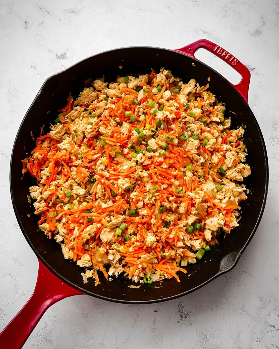 The image shows a black cast iron pan with a red handle, filled with a mix of cooked ingredients. The dish has three main layers visible: a base layer of small, crumbled light brown pieces that look like cooked turkey or chicken, mixed evenly with thin, bright orange shredded carrots scattered throughout. Small green bits, possibly chopped scallions or herbs, are sprinkled over the top, adding color contrast. The texture of the food looks soft and moist, with a slightly oily shine. The pan sits on a surface with a white marbled texture. photo taken with an iphone --ar 4:5 --v 7
