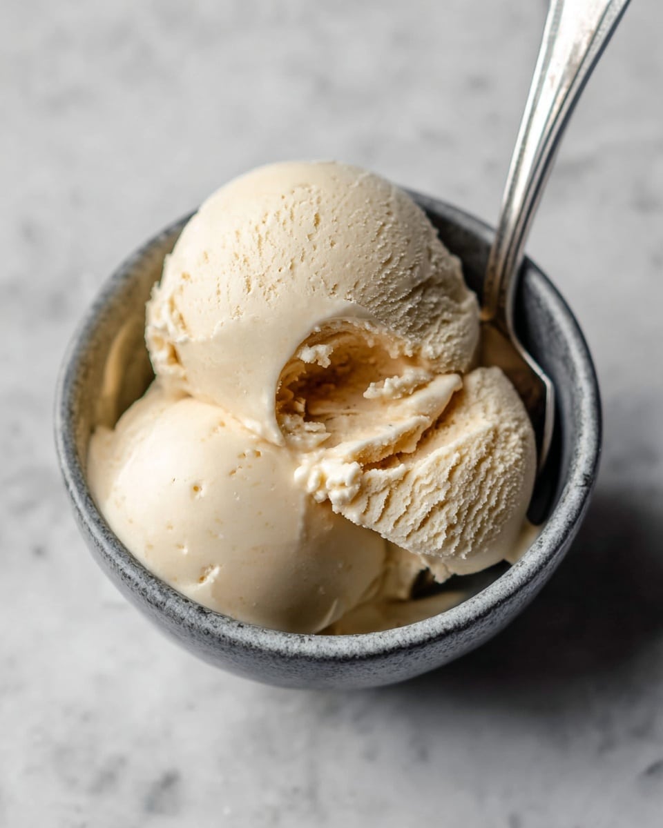 This image shows a small gray bowl filled with three scoops of light tan ice cream, which look creamy and smooth with a bit of softness around the edges. A silver spoon is placed inside the bowl, resting on the ice cream near the top. The bowl is set on a white marbled surface, adding a clean and simple background to the scene. The ice cream has a soft and rich texture, with the middle scoop having a bite taken out of it, showing off the dense inside. photo taken with an iphone --ar 4:5 --v 7