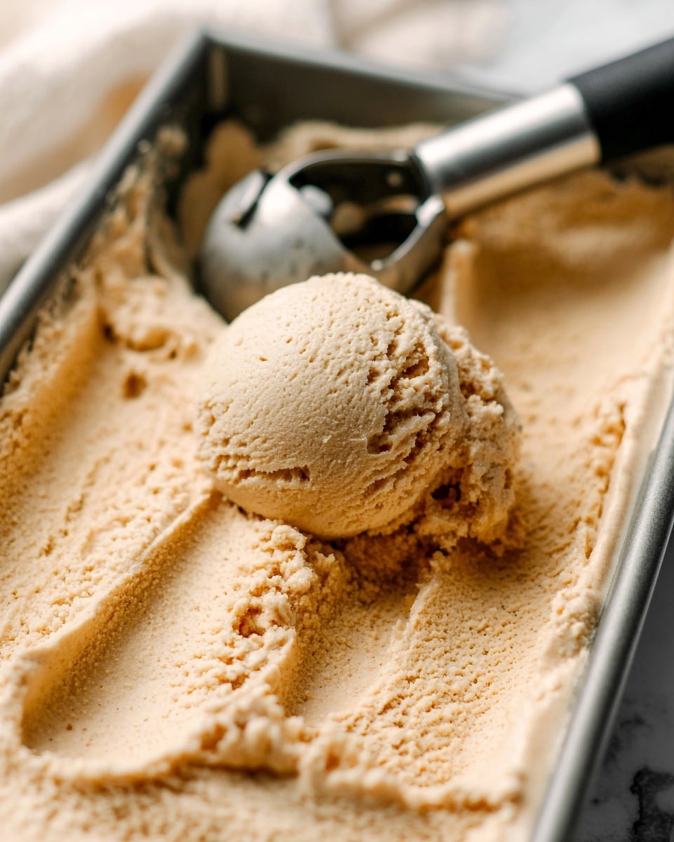 A close-up view of one large scoop of light brown ice cream with a creamy, slightly grainy texture placed in a rectangular metal container filled with the same ice cream. The ice cream has soft peaks and troughs, showing smooth movement from the scoop. A metal ice cream scooper with a black handle is partly embedded in the ice cream in the background. The surrounding gives a cozy, warm feel with soft, diffused light and a white marbled surface underneath. photo taken with an iphone --ar 4:5 --v 7