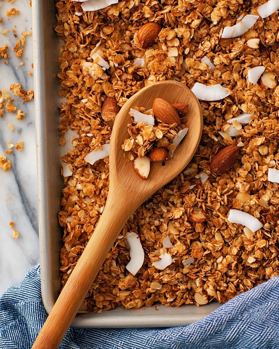 The image shows a close-up of crunchy granola scattered on a baking sheet. The granola has multiple layers of toasted oats in light and medium brown shades with a rough texture. There are white and light brown large, thin coconut flakes mixed evenly throughout. Whole almonds with a smooth, dark brown surface appear here and there, adding a varied color and texture. A wooden spoon with small holes filled with some granola rests on top, the spoon handle stretching diagonal across the image. The baking sheet edge is visible at the bottom, placed on a white marbled surface with a folded blue-striped cloth underneath. photo taken with an iphone --ar 4:5 --v 7