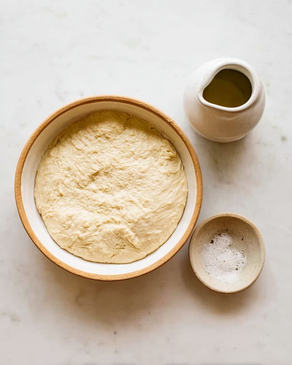 A white bowl with a light brown rim holds a single layer of pale beige dough that looks soft and slightly bubbly. To the right of the bowl, there is a small white ceramic jar with a spout filled with a greenish liquid, and below it, a tiny white ceramic bowl contains coarse salt. The items are placed on a white marbled surface with soft natural light showing clear textures and colors, photo taken with an iphone --ar 4:5 --v 7