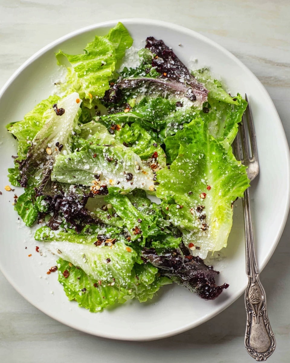 A white plate holds a fresh salad made of layers of green and dark purple lettuce leaves, each leaf showing crisp texture and natural veins. The lettuce is sprinkled with finely grated white cheese and small red chili flakes, adding contrast and detail. The salad sits on a white marbled surface. A silver fork with an ornate handle is resting slightly on the plate's edge. The overall look is fresh and simple, with the colors standing out clearly against the white plate and light background. photo taken with an iphone --ar 4:5 --v 7