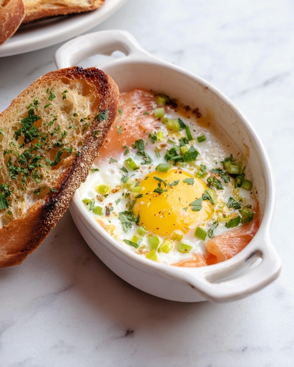 A white ceramic oval dish showing a cooked egg with a bright yellow yolk and white edges, topped with chopped green herbs and small green onion pieces. On one side, there is a piece of smoked salmon with a soft pink color, partially covered by the egg. A piece of toasted brown bread with visible air holes and sprinkled with green herbs leans against the side of the dish. The background is a white marbled surface. photo taken with an iphone --ar 4:5 --v 7