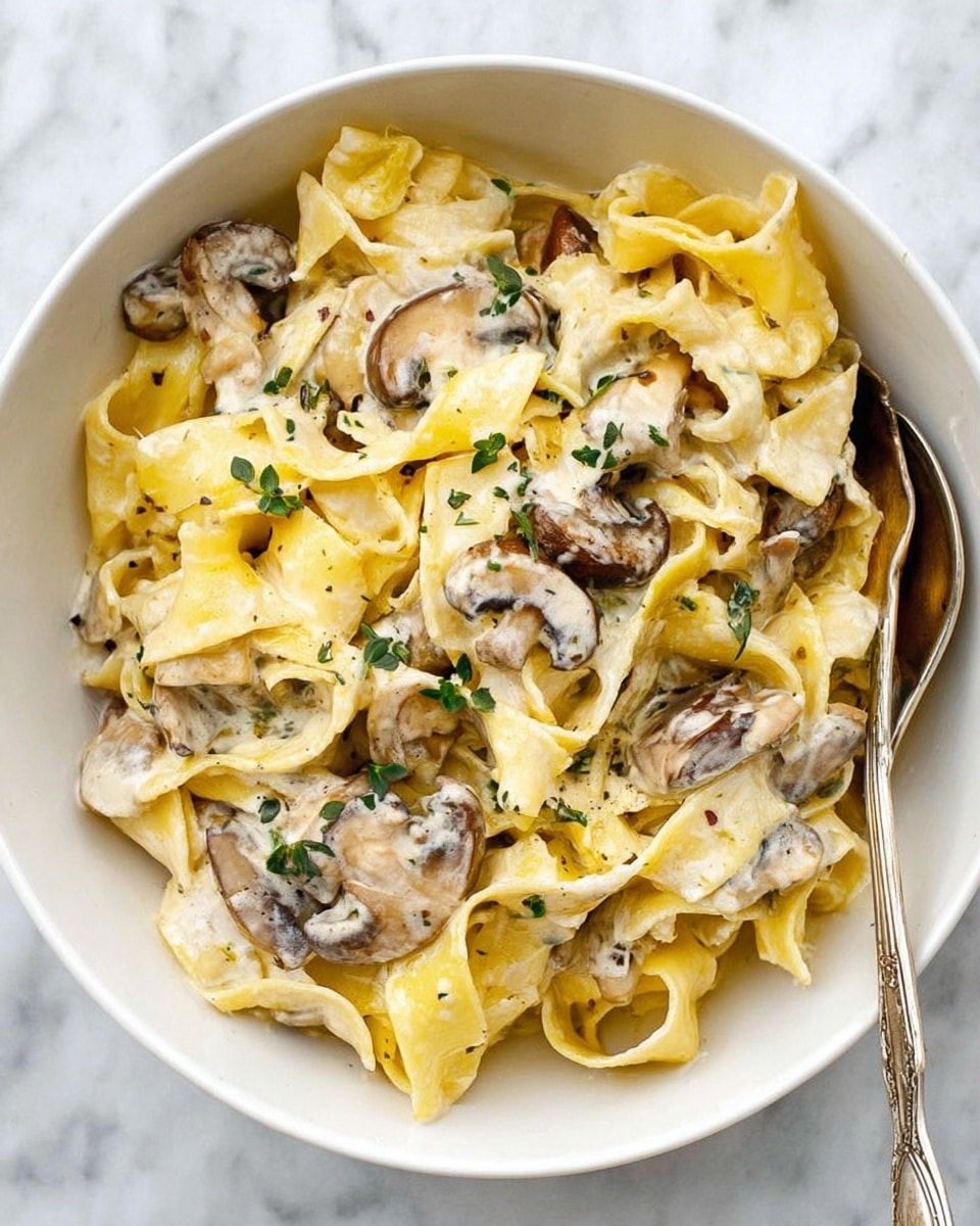 A white bowl filled with wide, flat yellow pasta noodles layered evenly with slices of cooked mushrooms that have a light brown color with darker edges, all mixed with a creamy white sauce coating every piece. Small green herb leaves are scattered lightly on top, adding small pops of color. A silver spoon rests in the bowl on the top right side. The bowl is placed on a white marbled surface. photo taken with an iphone --ar 4:5 --v 7
