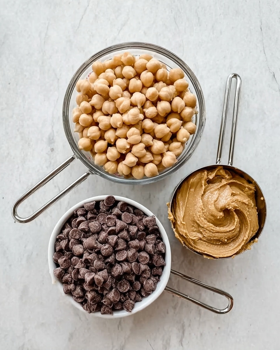 The image shows three measuring cups arranged on a white marbled surface. The top measuring cup is clear glass and filled with light beige chickpeas, showing their round shape clearly. Below it on the left is a white measuring cup filled with small, dark brown chocolate chips, with a slightly shiny texture. To its right is another white measuring cup filled with smooth, light brown peanut butter, showing a creamy texture with gentle swirls on the surface. All cups have metal handles pointing toward the right side of the image. photo taken with an iphone --ar 4:5 --v 7