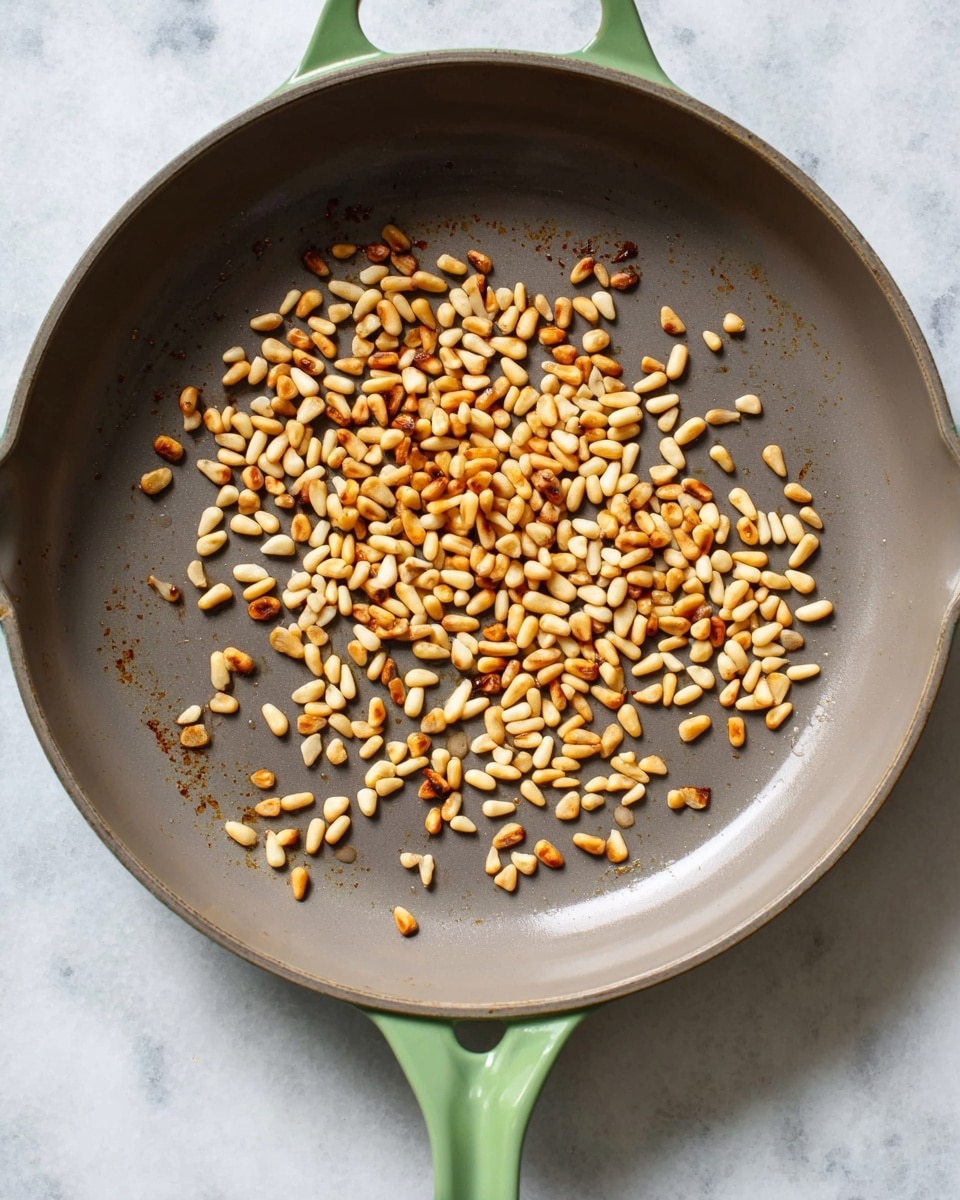 Inside a wide, shallow pan with a soft green handle, there is a single layer of small, slightly toasted pine nuts scattered mostly in one half of the pan. The pine nuts are golden brown with some lighter creamy-colored nuts mixed in, giving a toasted, crunchy look. The pan's interior is a smooth dark gray, contrasting with the light brown pine nuts, placed on a white marbled surface. photo taken with an iphone --ar 4:5 --v 7