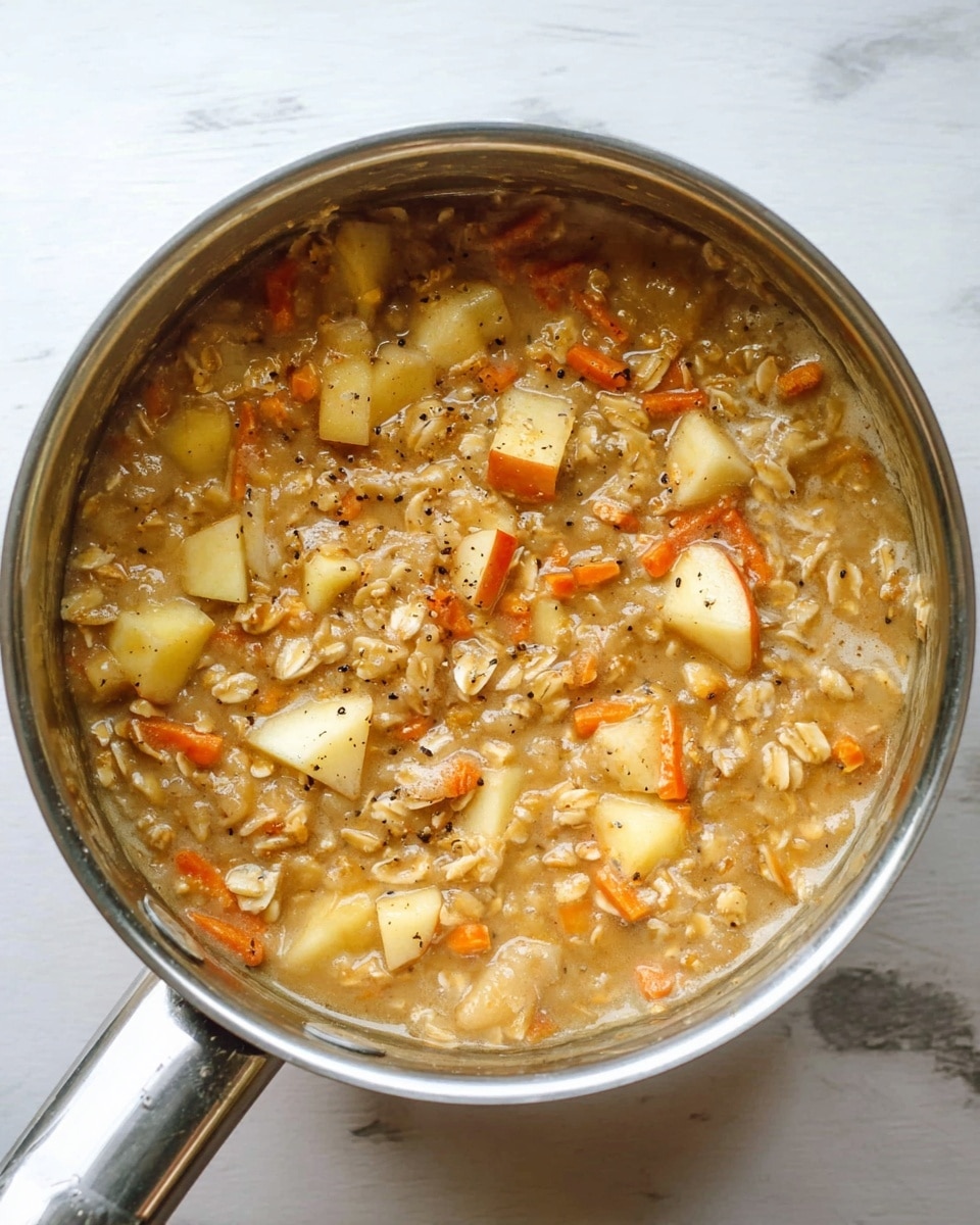 A close-up top view of a silver saucepan filled with a thick mixture of cooked oats, chunks of light yellow apples with skin, and small strips of orange carrot, all softly blended in a lightly brown, creamy liquid with visible black pepper specks, resting on a white marbled surface. photo taken with an iphone --ar 4:5 --v 7