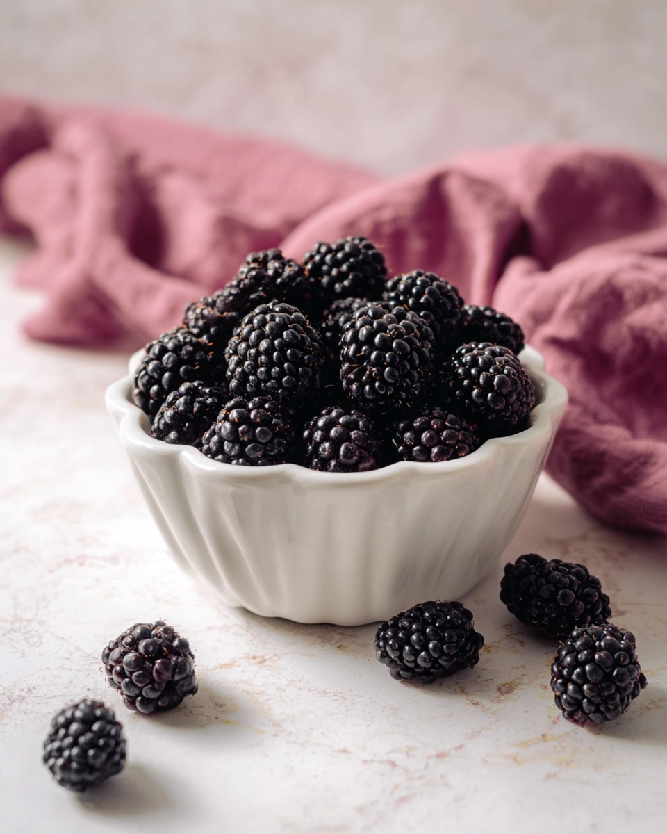 A white, fluted ceramic bowl filled with many shiny black blackberries, showing detailed texture and small round drupelets. Around the bowl, a few blackberries are scattered on a white marbled surface. In the background, a soft, folded pink cloth adds a splash of color. Photo taken with an iphone --ar 4:5 --v 7