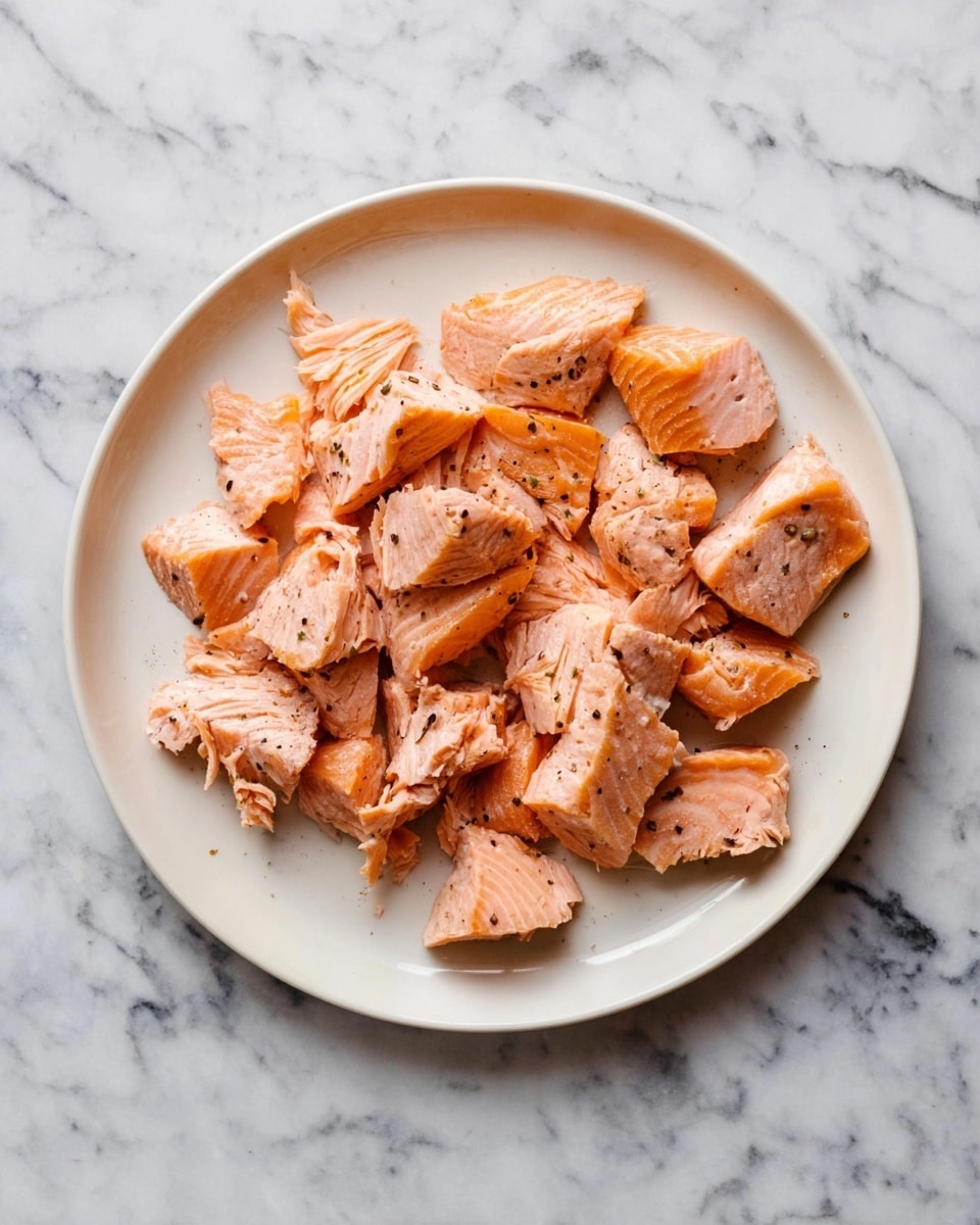 A white plate holds many pieces of cooked salmon scattered to fill the plate. The salmon is light pink with some darker orange edges and has a soft, flaky texture with visible layers in each piece. Small black pepper bits are sprinkled over the fish, adding contrast to the warm colors. The plate rests on a white marbled surface with gray veins, creating a clean and fresh look. photo taken with an iphone --ar 4:5 --v 7