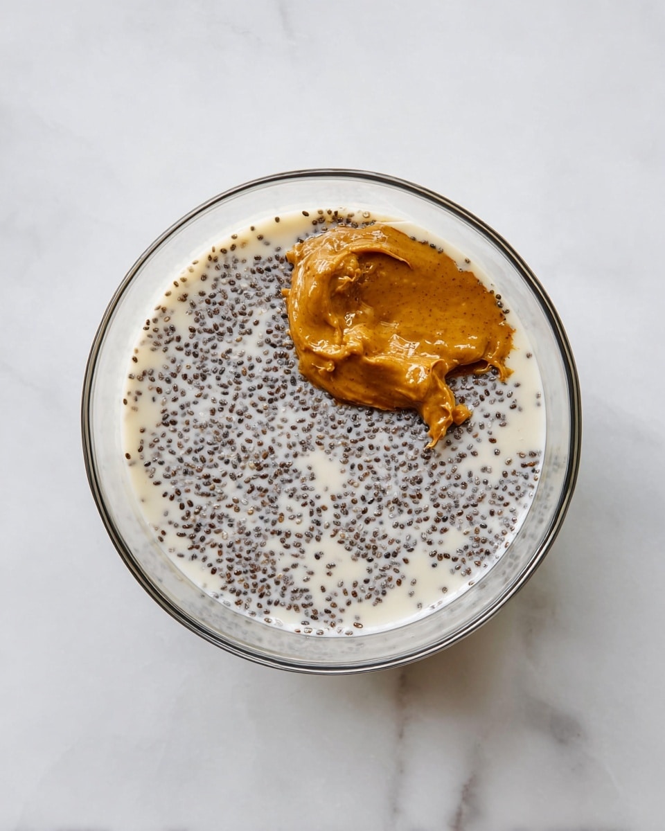 A clear glass bowl sits on a white marbled surface, containing a mixture of layers starting with creamy off-white milk at the base. Scattered throughout are small, round, dark chia seeds evenly spread in the milk layer. On top, near the center-right side, is a thick dollop of smooth, rich amber-colored nut butter. The layers show contrast between the pale milk, the tiny dark chia seeds scattered around, and the warm nut butter dollop. The photo is taken with an iphone --ar 4:5 --v 7
