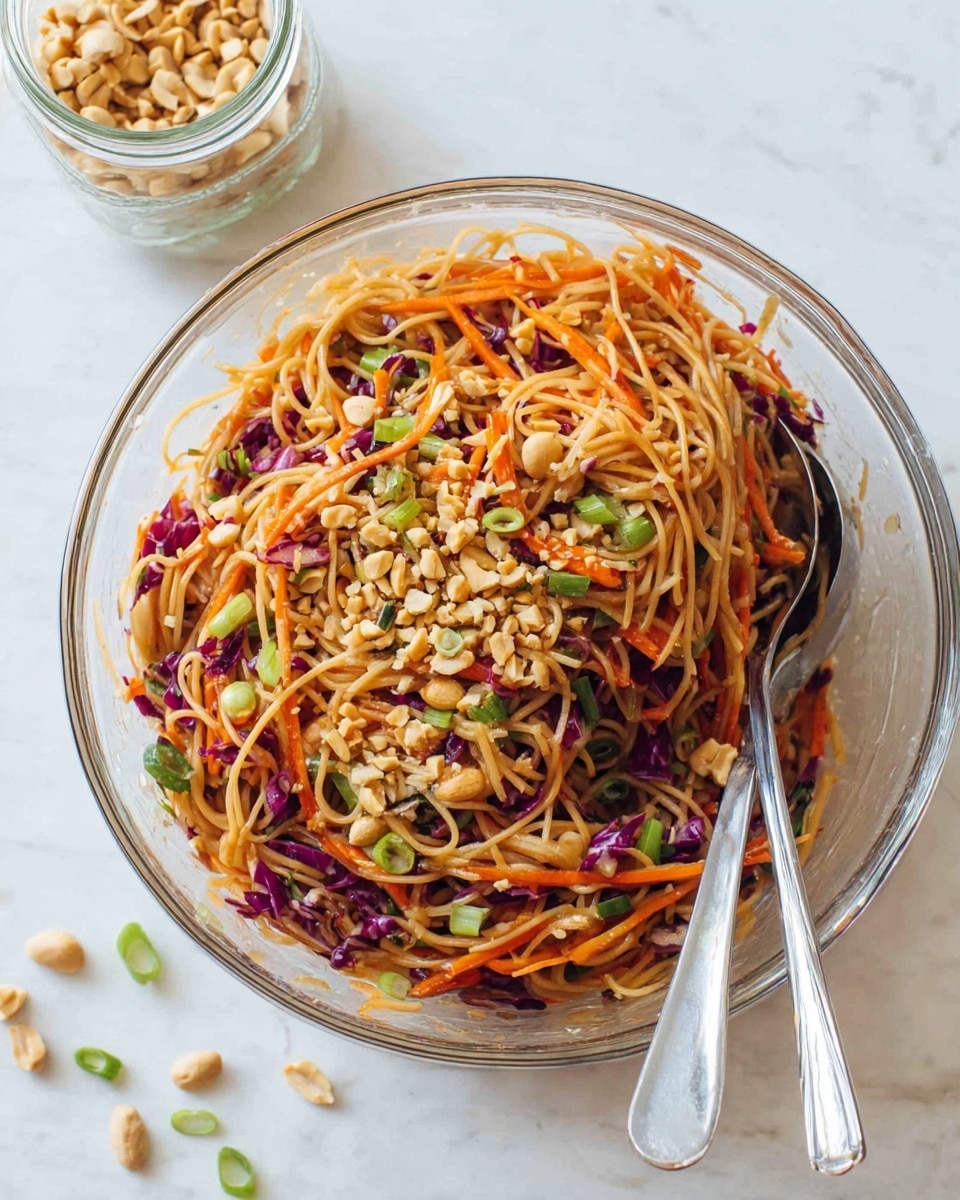 A large clear glass bowl filled with a mix of light brown noodles, thin orange carrot strips, small pieces of purple cabbage, and light green sliced scallions, all mixed together with crushed light tan peanuts sprinkled on top. Two shiny silver spoons rest inside the bowl on the right side. In the upper left corner, there is a blurry glass jar with more crushed peanuts. The bowl sits on a white marbled surface with a few scattered light green scallion rings nearby. Photo taken with an iphone --ar 4:5 --v 7