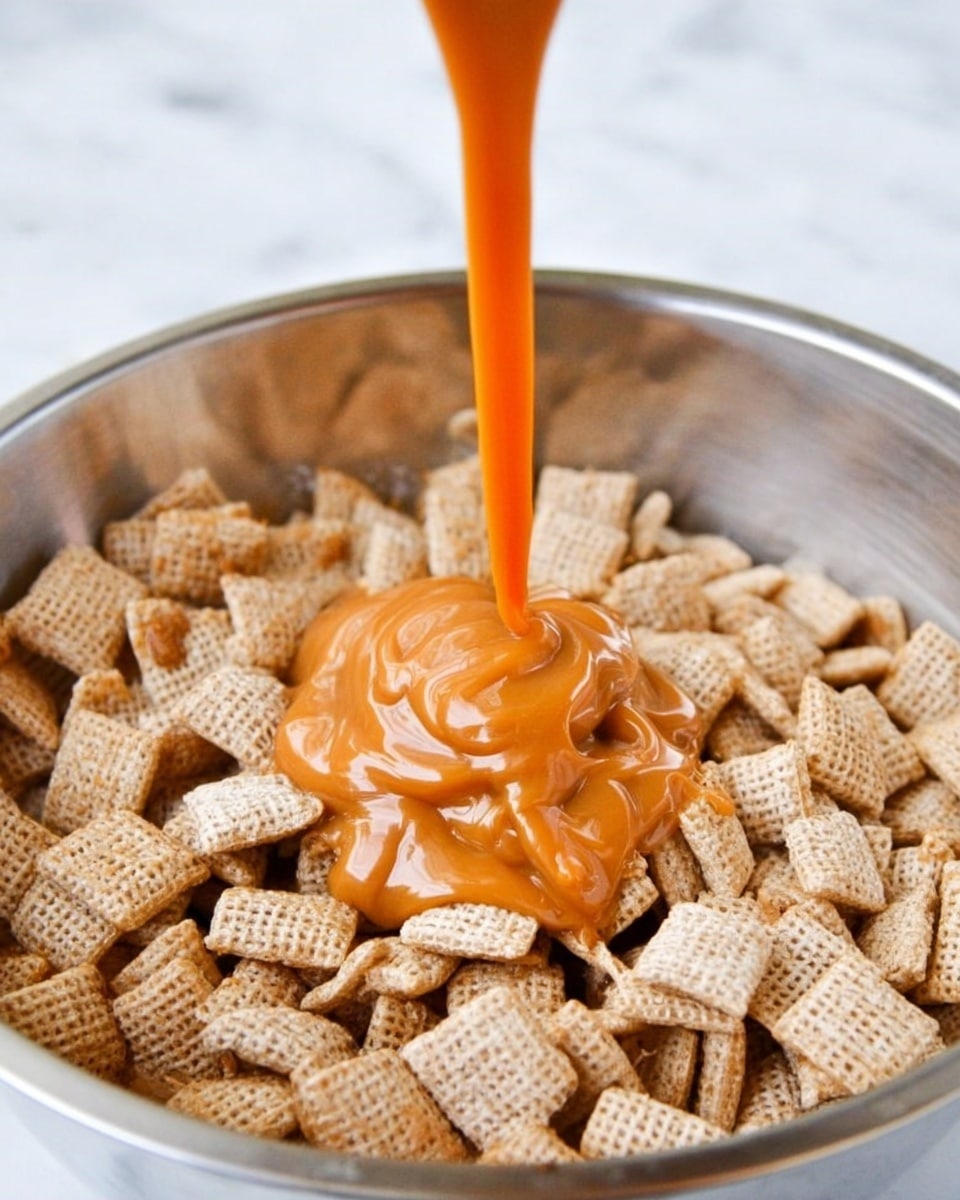 The image shows a silver bowl filled with many small square cereal pieces that are light brown and textured like woven fabric. Thick, smooth orange caramel sauce is being poured into the middle of the cereal, creating a soft mound on top of the cereal squares. The caramel has a shiny, sticky look and flows down slowly onto the cereal. The background is a white marbled texture. photo taken with an iphone --ar 4:5 --v 7