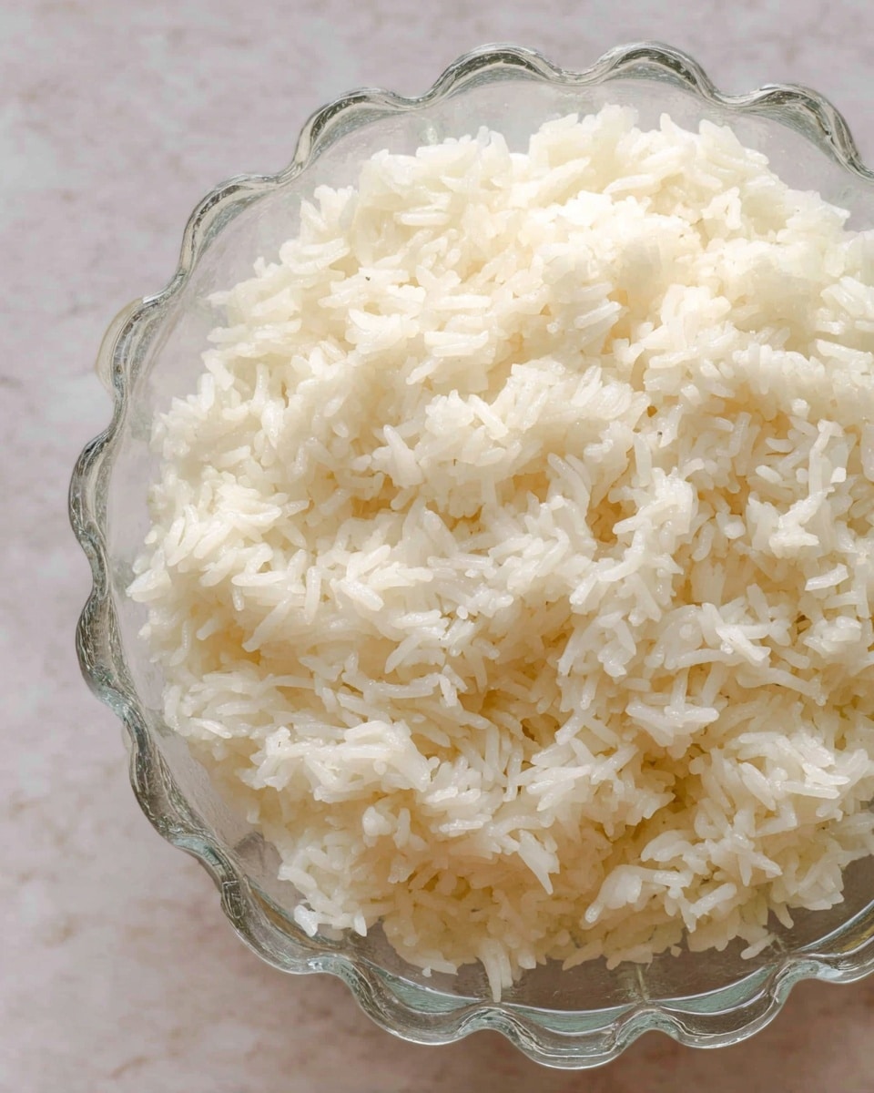 The image shows a close-up view of cooked white rice in a clear glass bowl with scalloped edges. The rice grains appear soft, slightly sticky, and have a shiny texture. The bowl is placed on a white marbled surface, and the photo focuses mainly on the rice's texture and the bowl's top half, with no other objects visible. photo taken with an iphone --ar 4:5 --v 7