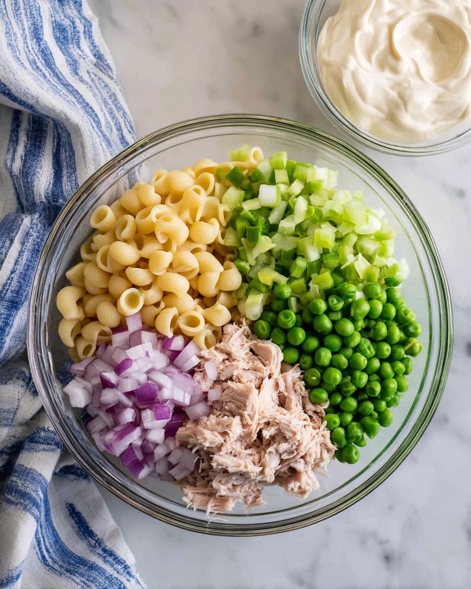 The image shows a clear glass bowl on a white marbled surface filled with five separate layers of ingredients: pale yellow small pasta shells at the top center, bright green peas to the right, light green chopped celery to the left, finely chopped purple and white onion in the center, and shredded light tan tuna at the bottom left. Next to this bowl is another smaller clear glass bowl filled with smooth, creamy white mayonnaise. A blue and white striped cloth napkin is partially visible in the top left corner. photo taken with an iphone --ar 4:5 --v 7