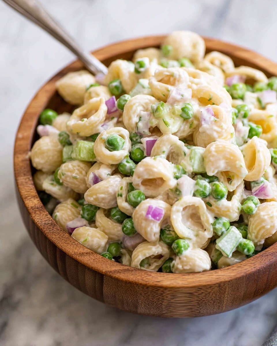 A close-up view of a creamy pasta salad served in a round wooden bowl placed on a white marbled surface. The salad consists of small shell pasta coated in a smooth, white dressing, mixed with bright green peas, small chunks of light green celery, and tiny pieces of purple onion scattered throughout. The textures look soft and fresh, with the peas adding a pop of color against the creamy pasta. A silver spoon is partially visible inside the bowl, ready to serve. photo taken with an iphone --ar 4:5 --v 7