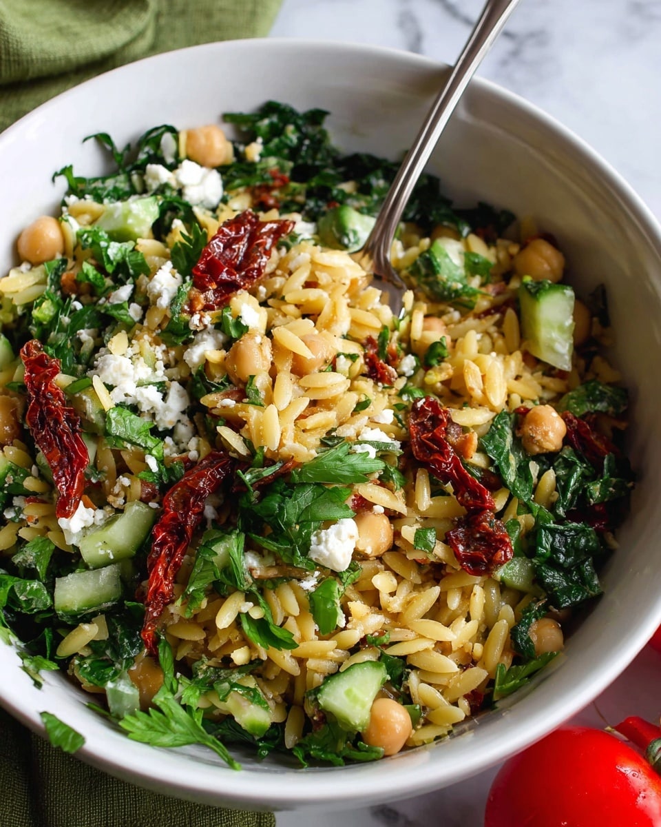 The image shows a white bowl filled with three main layers: the bottom layer is a mix of small, yellow orzo pasta, chickpeas, and chopped cucumber pieces, the middle layer contains dark green leafy greens and fresh parsley leaves scattered throughout, and the top layer is sprinkled with small crumbles of white cheese and thin strips of deep red sun-dried tomatoes. A metal fork is placed inside the bowl with some pasta, greens, and tomato pieces on it. The bowl sits on a white marbled surface with a green cloth partially visible nearby, and a red tomato is placed at the bottom right corner of the frame. photo taken with an iphone --ar 4:5 --v 7