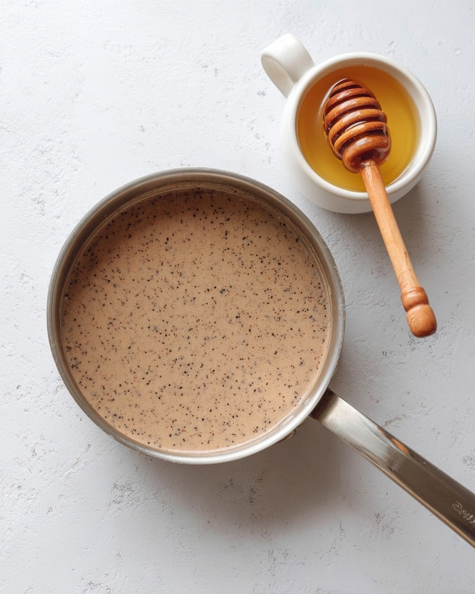 A top view of a silver saucepan filled with a light brown creamy liquid with tiny black seeds or specks throughout, placed on a white marbled surface. Next to it, a white cup holds honey with a wooden honey dipper resting inside, dripping some honey into the cup. photo taken with an iphone --ar 4:5 --v 7