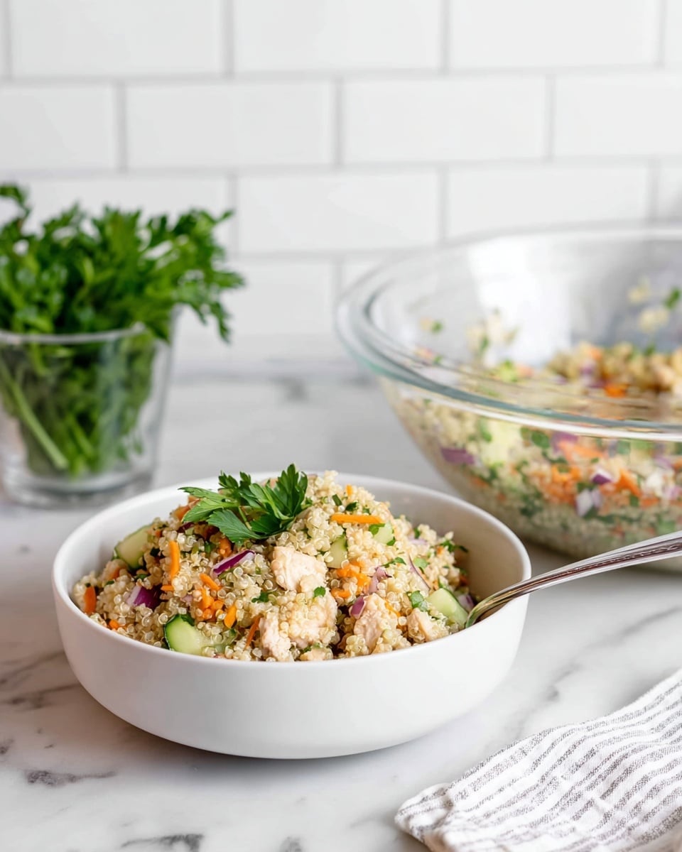 A white bowl filled with a colorful quinoa salad sits on a white marbled surface. The salad has several layers, starting with a base of fluffy, light beige quinoa mixed with small pieces of green cucumber, orange carrots, and red onion. There are also bits of light brown chicken mixed in, and a sprig of fresh green parsley on top for garnish. A silver spoon rests inside the bowl on the right side. Behind the bowl, a clear glass bowl shows more of the salad inside. To the left, a small clear glass holds bright green fresh parsley. The background features white subway tiles and a white striped cloth. Photo taken with an iphone --ar 4:5 --v 7