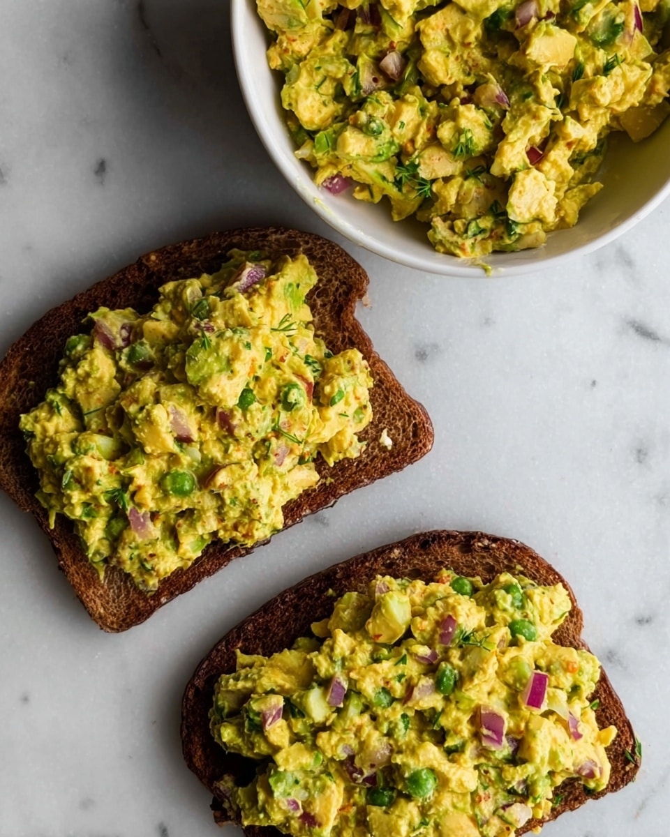 The image shows two pieces of dark toasted bread placed side by side on a white marbled surface. Each toast is topped with a thick layer of a mixed yellow and green salad spread that contains visible small chunks of red onion and green herbs throughout. Above the toasts, there is a white bowl filled with the same chunky yellow salad mixture, featuring uneven textures and small pieces of vegetables visible. The colors are warm with a focus on yellow, green, and bits of red, all laid out simply and naturally. Photo taken with an iphone --ar 4:5 --v 7