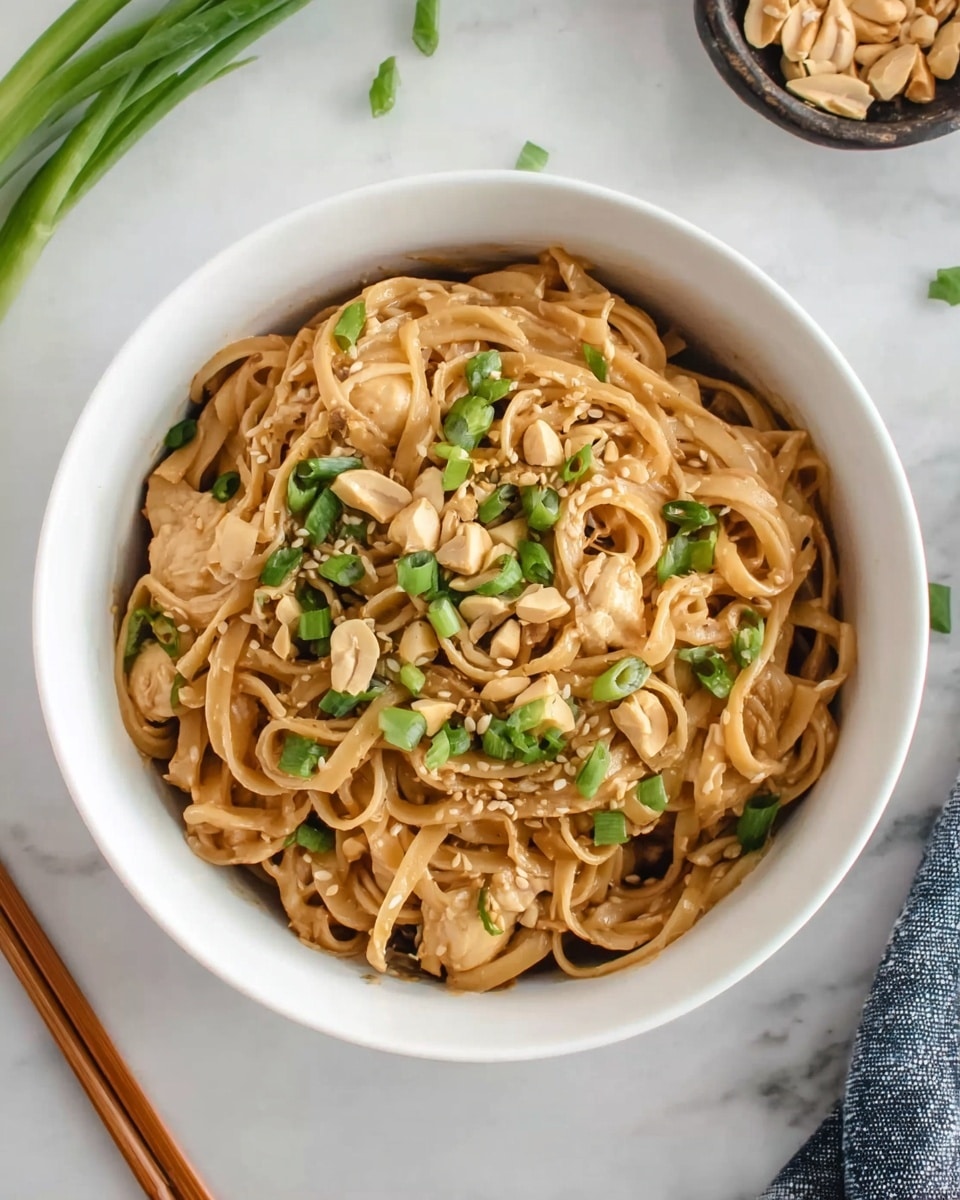 A white bowl filled with a swirl of brown noodles, mixed with light tan pieces of chicken, topped with chopped green onions and sprinkled with light brown peanuts and sesame seeds. The bowl sits on a white marbled surface, with some green onion stalks and chopsticks nearby, adding more color to the scene. Photo taken with an iphone --ar 4:5 --v 7