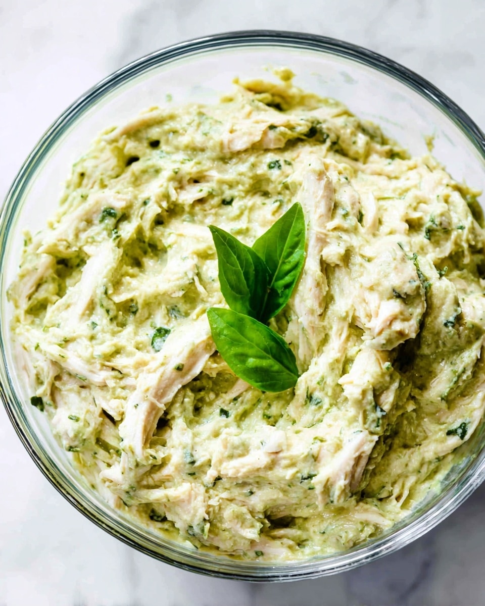 A close-up view of a clear glass bowl filled with a creamy, pale green mixture that has a thick, slightly chunky texture. Within the mixture, shredded pieces of white chicken are visible, mixed evenly throughout. On top, there are two fresh green basil leaves adding a pop of color. The bowl is set on a white marbled surface that contrasts gently with the green tones of the dish. photo taken with an iphone --ar 4:5 --v 7