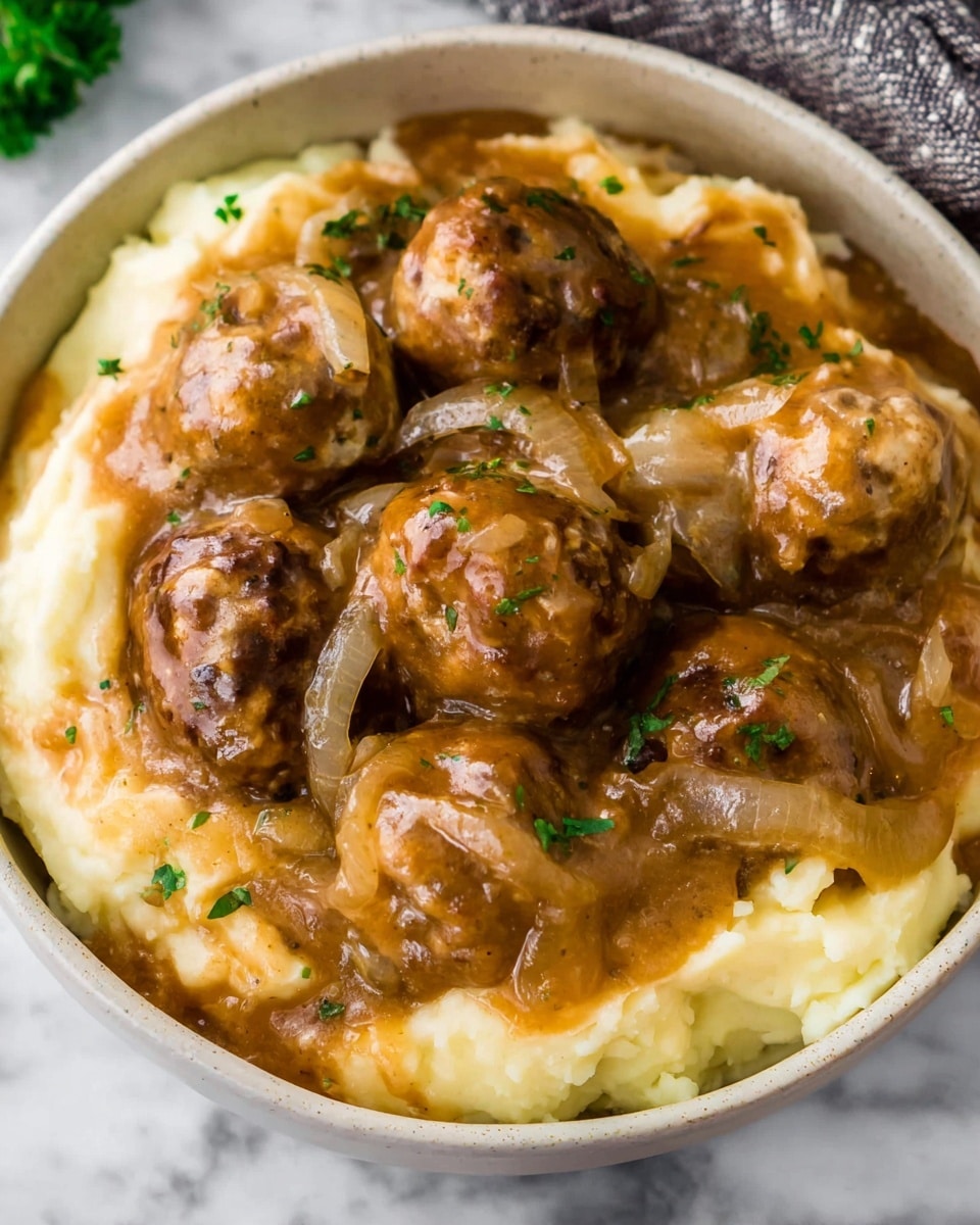 A white bowl filled with two main layers: the bottom layer is smooth mashed potatoes with a creamy pale yellow color, and the top layer has several brown meatballs covered in a thick brown gravy with visible slices of cooked onions mixed in. The gravy looks glossy and coats the meatballs well, adding a rich texture. Small bits of green herbs are scattered lightly over the dish. The bowl sits on a white marbled surface. photo taken with an iphone --ar 4:5 --v 7