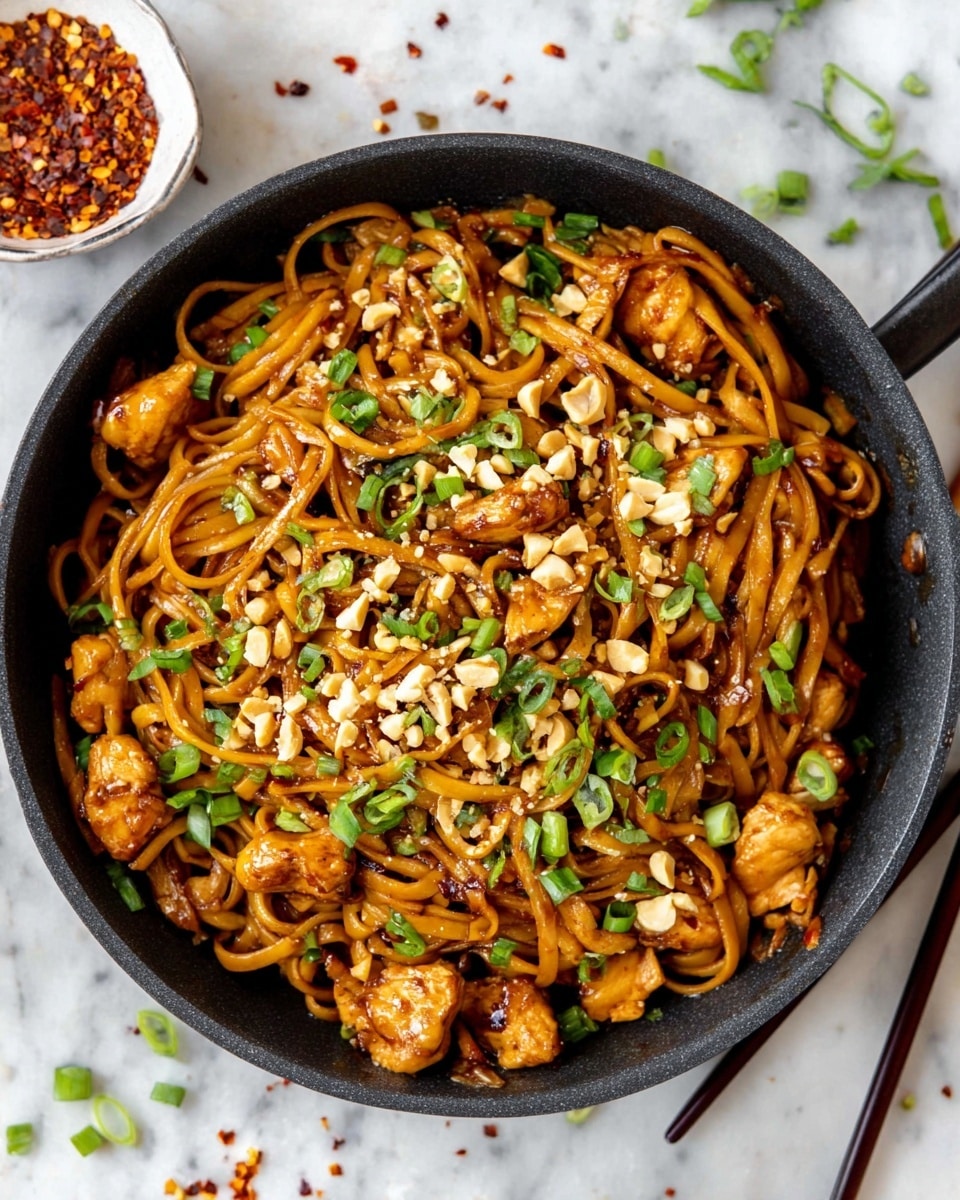 A black pan filled with glossy brown noodles mixed with small chunks of golden brown cooked chicken, topped with bright green chopped scallions and scattered crushed peanuts. The noodles and chicken have a shiny, slightly sticky sauce coating. Around the pan on a white marbled surface are some scattered red chili flakes and chopped green onions, along with black chopsticks lying on the right side. Photo taken with an iphone --ar 4:5 --v 7
