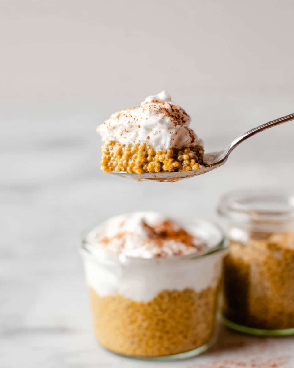 The image shows a close-up of a spoon holding a small portion of chia seed pudding a textured golden brown bottom layer that looks soft and moist, and a fluffy white cream layer on top, dusted lightly with a brown powder. Below the spoon, two small glass jars are filled with the same dessert, with the golden bottom layer filling most of the jars and a thick white cream layer on top, sprinkled with the same brown powder. The jars are set on a white marbled surface with a clean, soft background. photo taken with an iphone --ar 4:5 --v 7