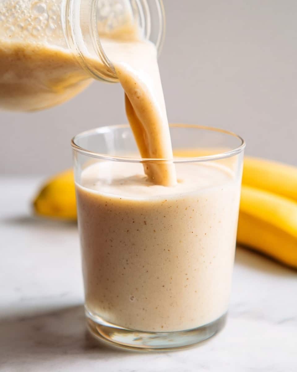A close-up view of a thick, creamy beige smoothie being poured from a clear glass bottle into a clear glass cup. The smoothie has a smooth texture with tiny specks visible throughout, showing its natural ingredients. The glass cup is nearly full, with fresh bubbles forming on the top layer. In the background, a whole ripe banana with bright yellow skin is partly visible, resting on a white marbled surface. The image has soft, natural lighting highlighting the smoothie's rich color and freshness, photo taken with an iphone --ar 4:5 --v 7