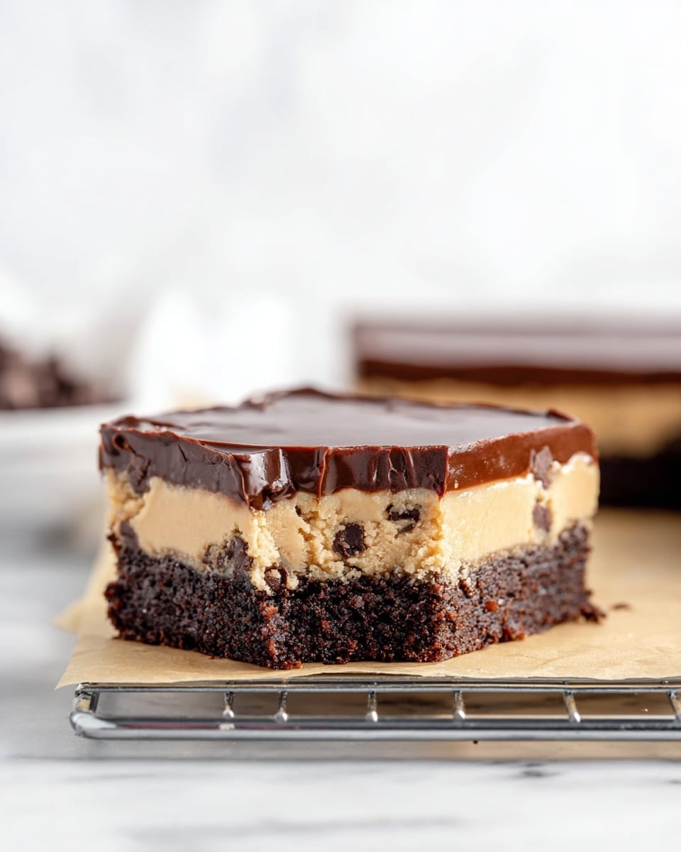 A thick square dessert bar sits on a metal rack over parchment paper, placed on a white marbled surface. The dessert has three clear layers: the bottom is a dense, dark brown brownie with a rough texture, the middle layer is a smooth, creamy light tan cookie dough mixed with chocolate chips, and the top is a thin, shiny dark chocolate ganache layer, all evenly stacked. The background is softly blurred white making the dessert the clear focus. photo taken with an iphone --ar 4:5 --v 7
