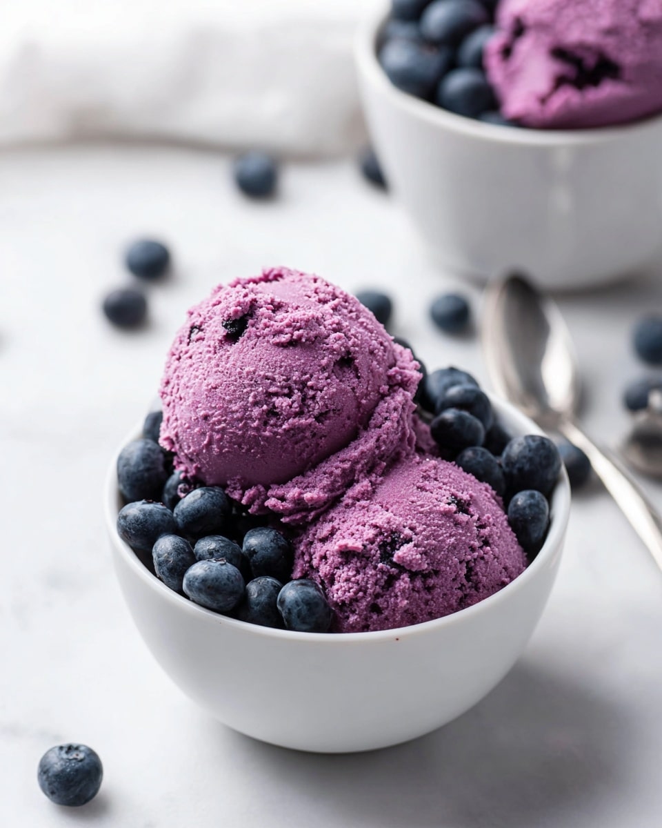 A white bowl holds two scoops of bright purple blueberry mousse with a light, slightly chunky texture on top, sitting next to a small pile of fresh, plump dark blue blueberries inside the same bowl. In the background, there is another white bowl filled with more blueberries, and a few loose blueberries are scattered on the white marbled surface around the bowls. The setting is bright and clean with soft natural light. Photo taken with an iphone --ar 4:5 --v 7