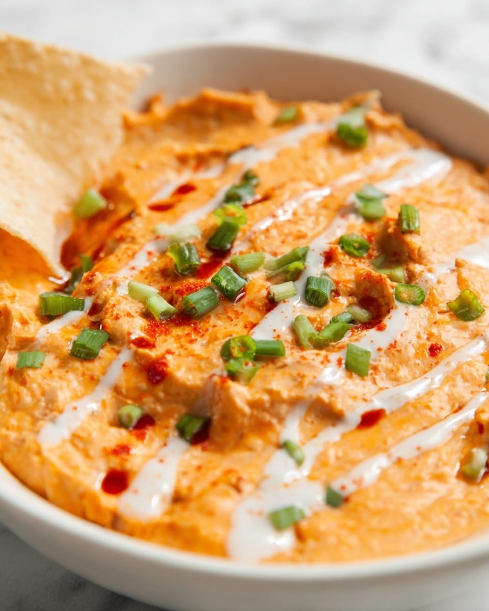 The image shows a close-up of a creamy orange dip with a smooth and slightly chunky texture spread inside a white bowl placed on a white marbled surface. The dip has small green onion pieces scattered on top, adding bright green color spots, and thin white sauce lines drizzled across it. There are some red hot sauce drops providing contrast. A crisp, light brown chip is partially dipped into the bowl near the left side, adding a crunchy element. The lighting gives the dip a fresh and appetizing look. Photo taken with an iphone --ar 4:5 --v 7