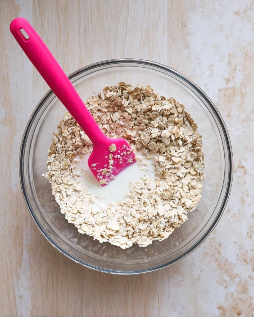 A clear glass bowl filled with dry oatmeal flakes covering most of the bowl, with a small pool of white liquid, likely milk, visible near the center. A bright pink spatula rests inside the bowl, partially submerged in the oatmeal and liquid mix, positioned slightly diagonally with the handle extending out of the bowl. The bowl is set on a white marbled textured surface. photo taken with an iphone --ar 4:5 --v 7