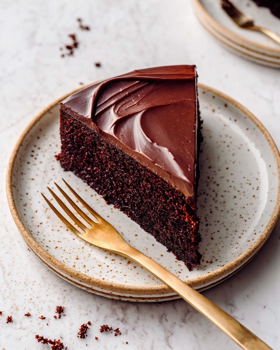 A single slice of dark chocolate cake sits on a white plate with small brown speckles and a textured rim. The cake has two layers: a thick, moist, dark brown base layer with a crumbly texture, topped by a smooth, glossy layer of chocolate ganache with gentle swirls. A gold fork rests on the plate, pointing to the cake slice. The background is a white marbled surface with scattered crumbs near the plate. Photo taken with an iphone --ar 4:5 --v 7
