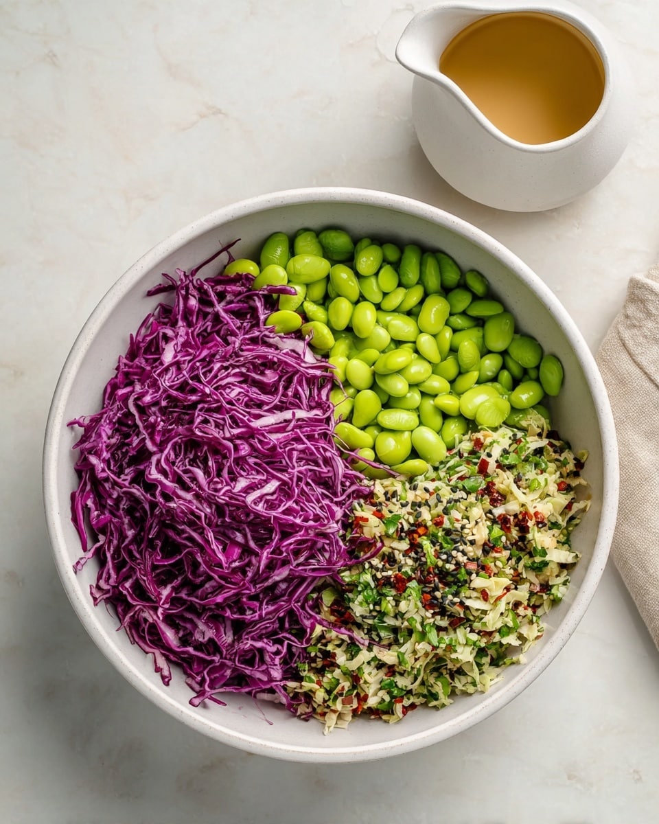 A white bowl sits on a white marbled surface, filled with three distinct layers. On the left side is a pile of thinly shredded purple cabbage, its vibrant color and crisp texture prominent. To the right of the cabbage, bright green edamame beans take up about a third of the bowl, showing a smooth and shiny surface. The last section, at the bottom right, is a mix of finely chopped ingredients including white cabbage, green herbs, small red pepper pieces, and black sesame seeds, adding texture and color contrast. Nearby, on the right side of the image, a small white pitcher holds a light brown dressing. photo taken with an iphone --ar 4:5 --v 7