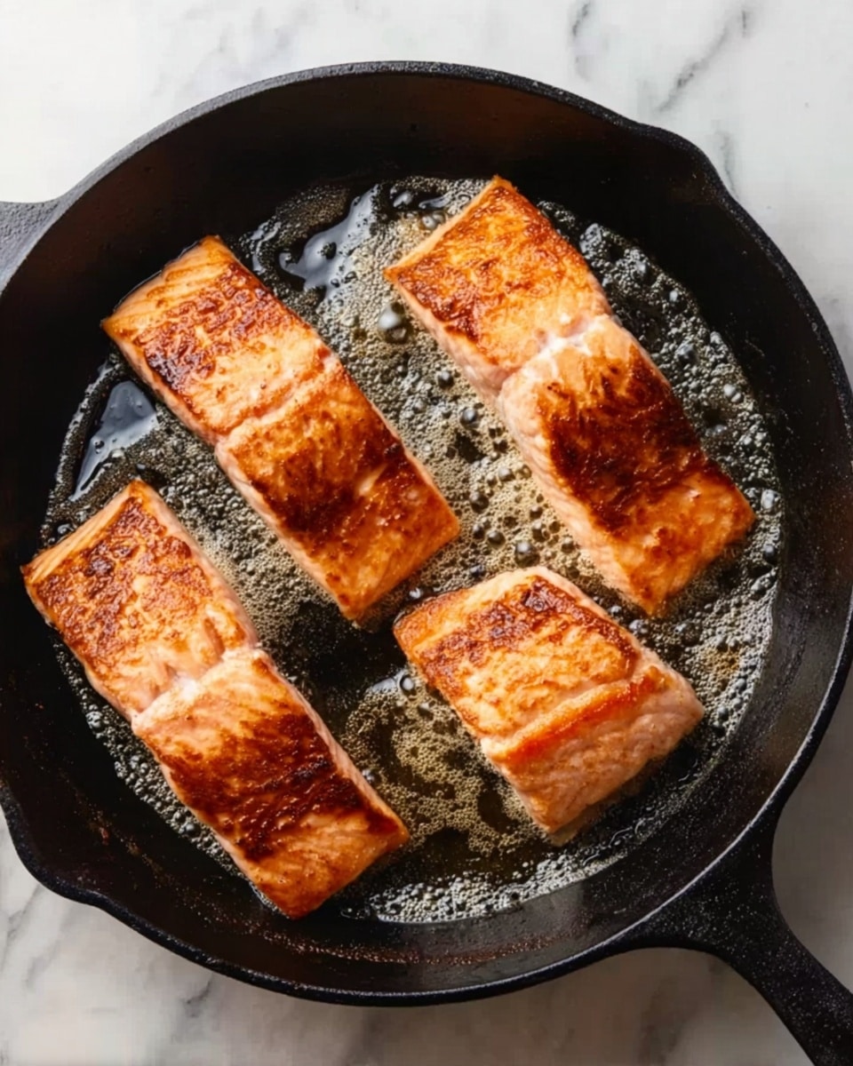 The image shows four pieces of salmon cooking in a black cast iron pan on a white marbled surface. Each salmon piece is thick and rectangular with crispy golden-brown skin facing up, showing a slightly rough texture with a few browned edges. The pan has shiny melted butter bubbling around the salmon, adding a glossy look to the cooking surface. The pieces are arranged evenly to cover the bottom of the pan, and the salmon's colors shift from light pink to golden brown where it is cooked. Photo taken with an iphone --ar 4:5 --v 7